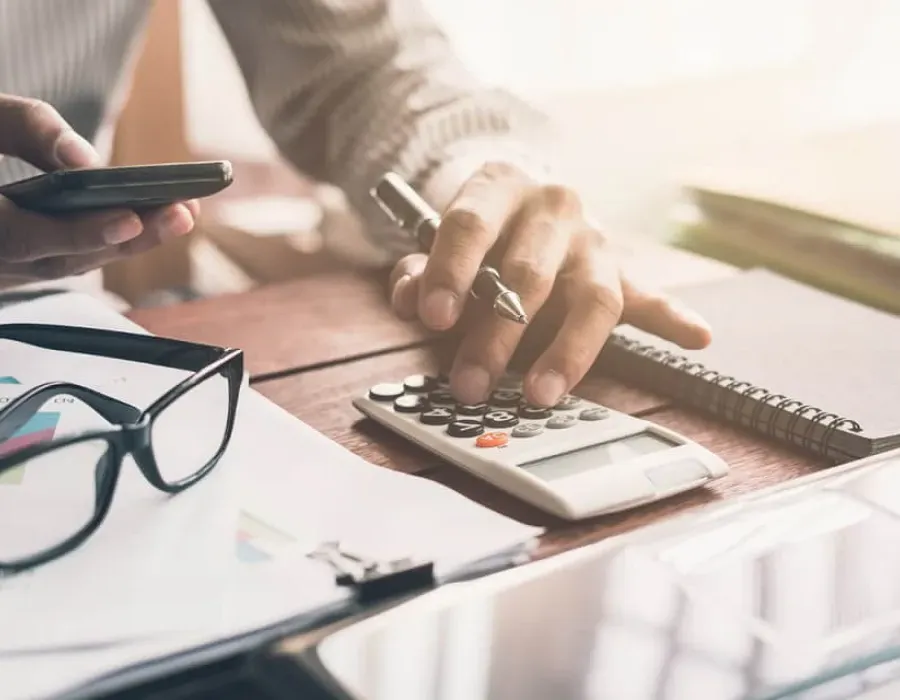 Accountant punching in calculations on calculator with glasses resting on desktop